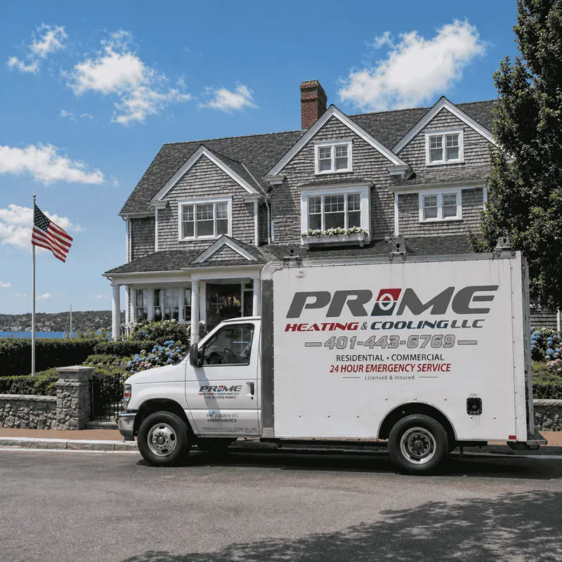 A white Prime Heating and Cooling service truck parked in front of a large Newport, RI home with gray shingles, white trim, and landscaped gardens on a sunny day.